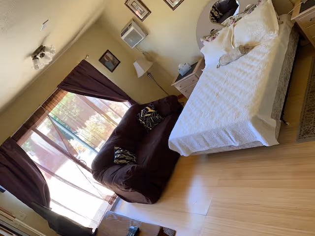 Bright bedroom with a bed, a brown loveseat, nightstands and a large window with curtains.