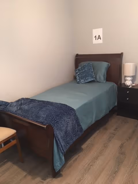A small bedroom with a single wooden bed made with teal bedding and a dark blue throw blanket. There is a matching teal pillow and a decorative blue pillow on the bed. Next to the bed is a black nightstand with a white lamp and some items on it. A wooden chair is partially visible on the left side. The wall above the bed has a paper sign labeled '1A'. The floor is wood with a light brown finish.