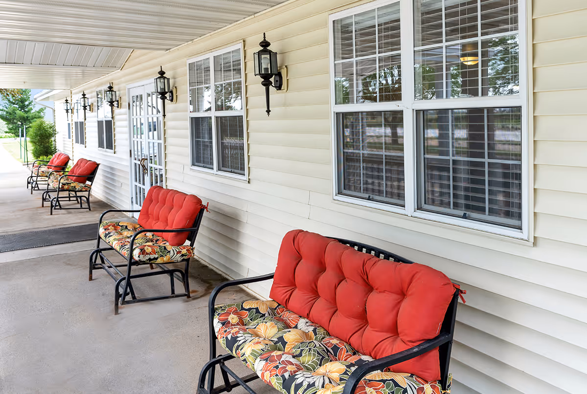 Covered outdoor patio area with several black metal benches featuring red and floral cushions lined up against the cream-colored siding of a building with multiple windows and black lantern-style wall lights.