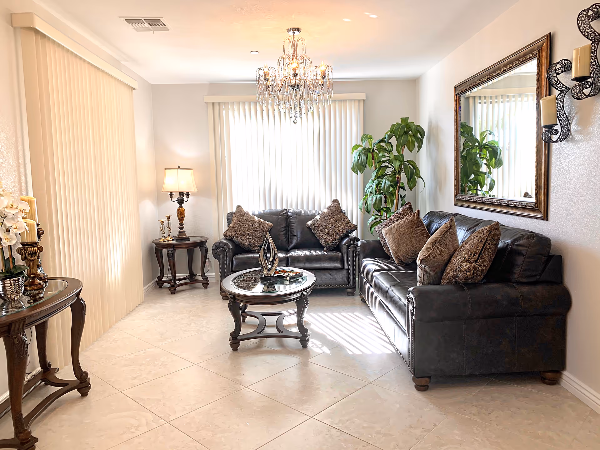 A bright living room with two dark leather sofas adorned with multiple decorative pillows. A round glass-top coffee table with a decorative centerpiece sits in the middle. There is a large mirror on the wall above one sofa, a tall green plant in the corner, and a side table with a lamp and small decor items. Vertical blinds cover the windows, and a chandelier hangs from the ceiling. The floor is tiled with light-colored tiles.