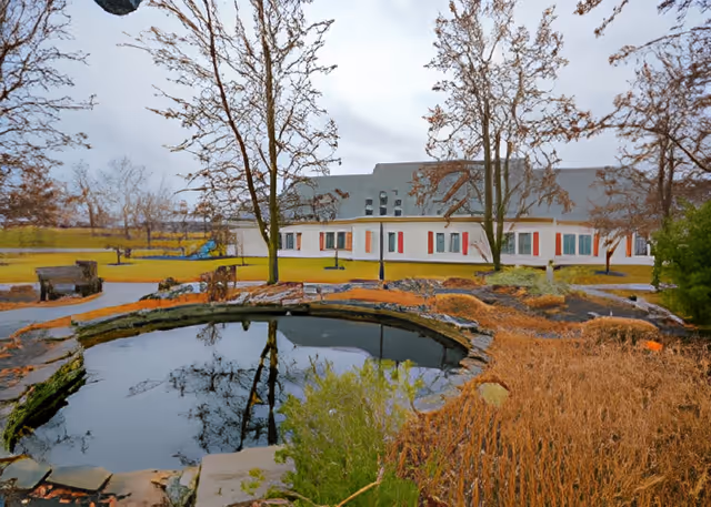 A small landscaped pond and leafless trees in front of a low, light-colored building on an overcast day.