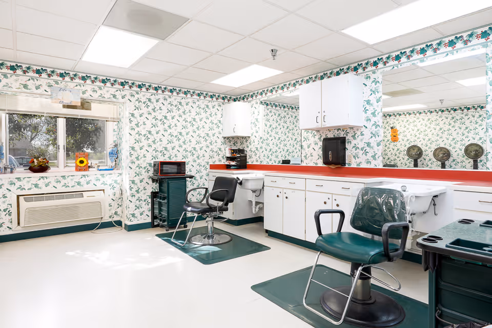 Interior of a salon room in an assisted living facility with two salon chairs, sinks for hair washing, white cabinets with red countertops, floral wallpaper, a window with blinds, and a microwave on a small cart.