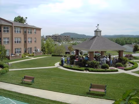 Grassy courtyard with benches and a gazebo where a group of people are gathered beside a brick retirement building and distant hills.