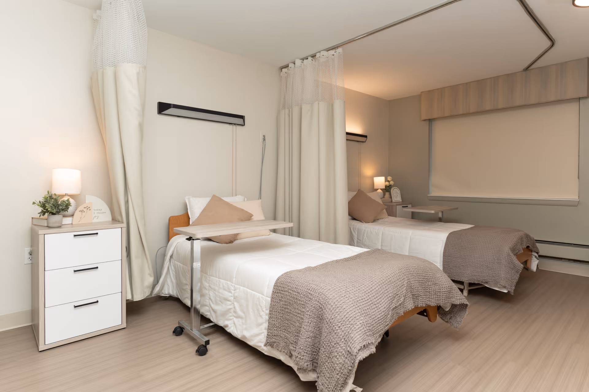 A clean and well-lit room with two single beds separated by a privacy curtain. Each bed has white bedding, a beige throw blanket, and a beige pillow. There are bedside tables with lamps and small decorative items next to each bed. The room has light-colored walls and flooring, with a large window covered by a beige roller blind.