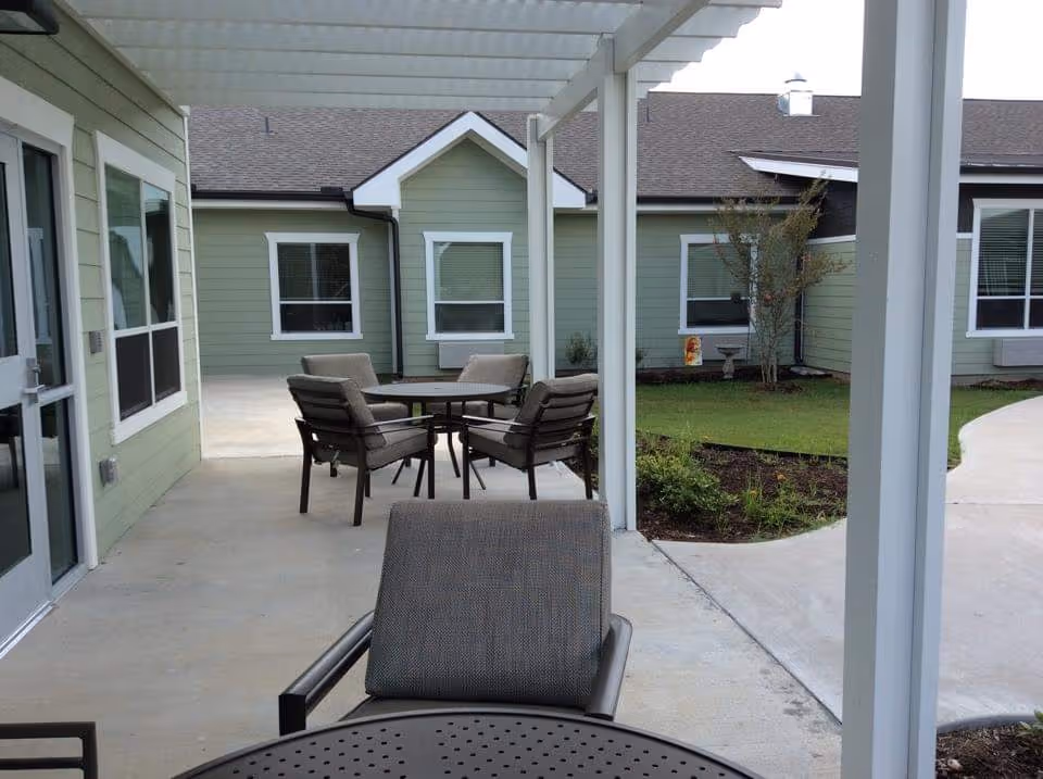 Outdoor patio area at Arbor Hills Rehabilitation and Healthcare Center with cushioned chairs and round tables under a white pergola. The patio is adjacent to a green building with white-trimmed windows and a small landscaped garden with grass and plants.