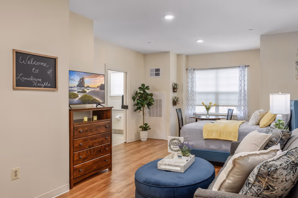 A cozy senior living facility room at Lansdowne Heights featuring a bed with yellow and gray bedding, a small round dining table with two chairs by a window with patterned curtains, a wooden dresser with a TV on top, a potted plant, and a comfortable seating area with a blue ottoman and a couch with multiple pillows. A chalkboard on the wall reads 'Welcome to Lansdowne Heights.' The room has wood flooring and soft lighting.