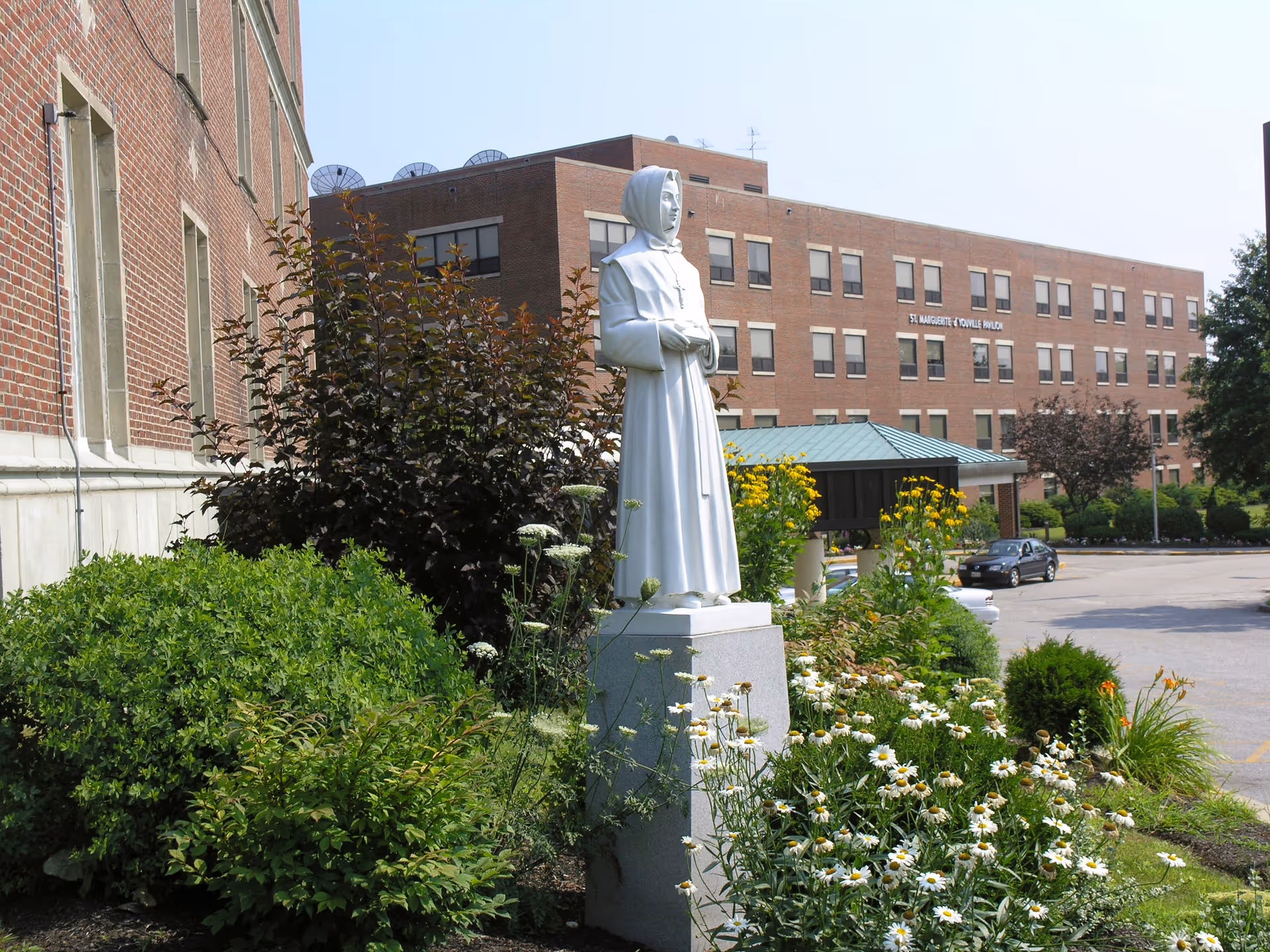 A white statue of a nun stands among flowers and shrubs in front of a brick senior living building entrance.