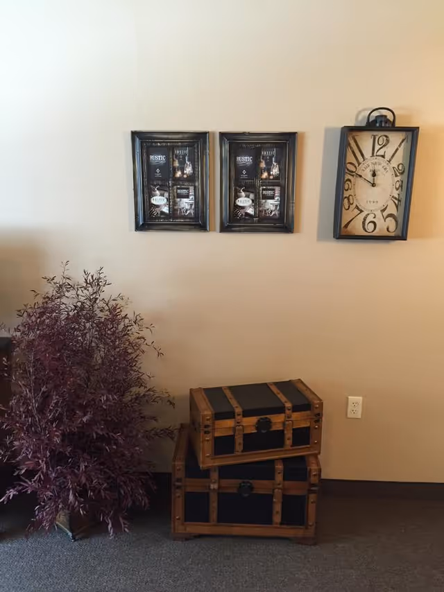 A beige wall with two framed rustic-themed pictures and a vintage-style rectangular clock hanging above two stacked wooden trunks with black panels. To the left of the trunks is a potted plant with dark red leaves. There is a power outlet on the wall near the floor.