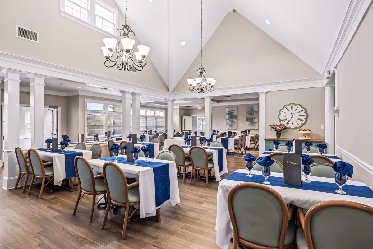 A bright and spacious dining room with multiple tables covered in white tablecloths and blue runners. Each table is set with blue napkins folded in glasses and menus. The room features high vaulted ceilings with chandeliers, large windows allowing natural light, and decorative wall art and a large clock on the walls.