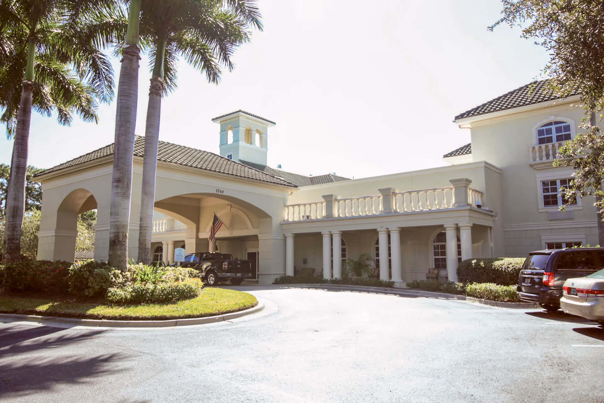 Entrance and porte-cochère of a cream-colored senior living building with palm trees and parked cars.
