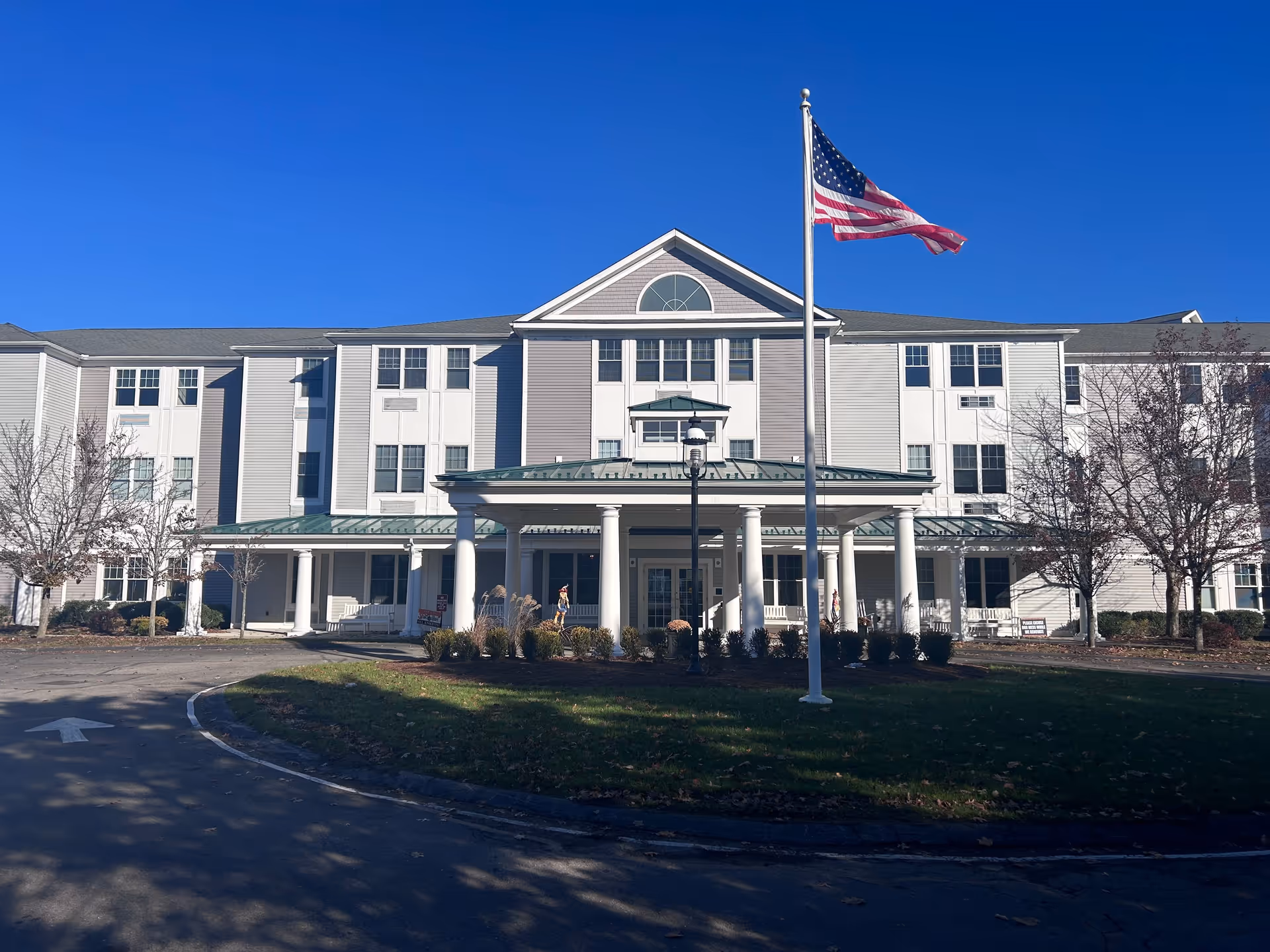 Front exterior view of a three-story senior living facility building with white and gray siding, a covered entrance supported by white columns, an American flag on a flagpole in front, and leafless trees on either side under a clear blue sky.