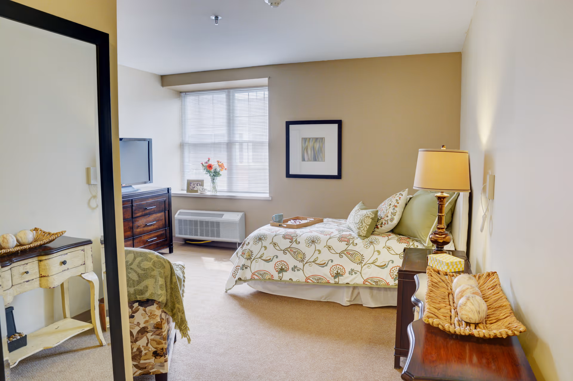 A cozy bedroom with a bed covered in floral patterned bedding and multiple pillows. There is a wooden nightstand with a lamp and a decorative basket on it. A dresser with a flat-screen TV on top is positioned near a window with blinds. A framed abstract artwork hangs on the wall above the bed. A large mirror reflects part of the room, including a small table with decorative items and a chair with a green throw blanket.
