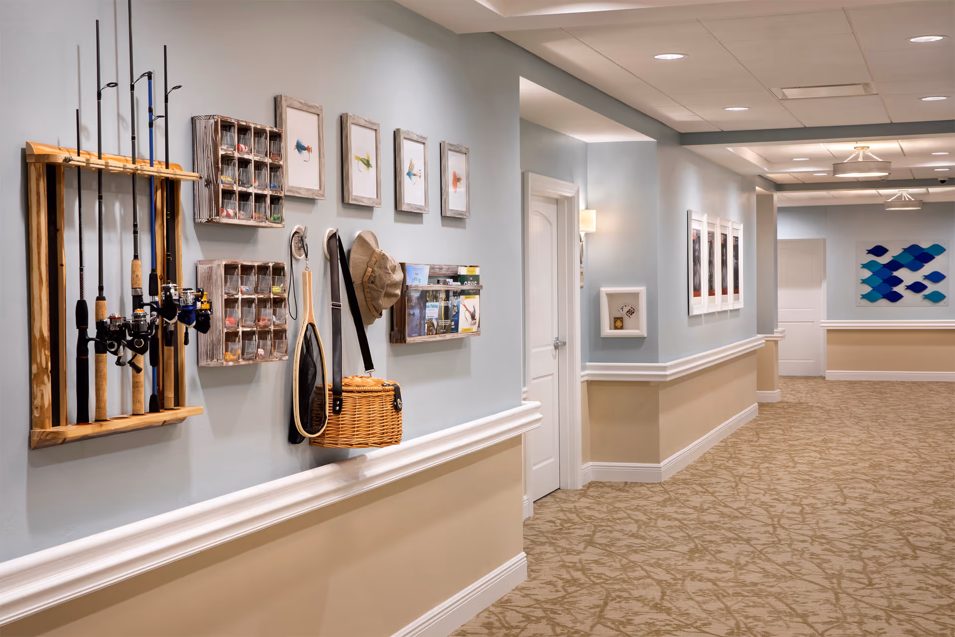 A well-lit hallway in a senior living facility with beige patterned carpet and light blue and beige walls. On the left wall, there are fishing rods mounted on a wooden rack, small wooden shelves holding fishing lures, a hat, a fishing net, and a wicker basket. Several framed pictures and brochures are also displayed on the wall. The hallway has white doors and decorative wall moldings, with additional framed artwork and a blue fish-themed wall decoration visible further down the corridor.