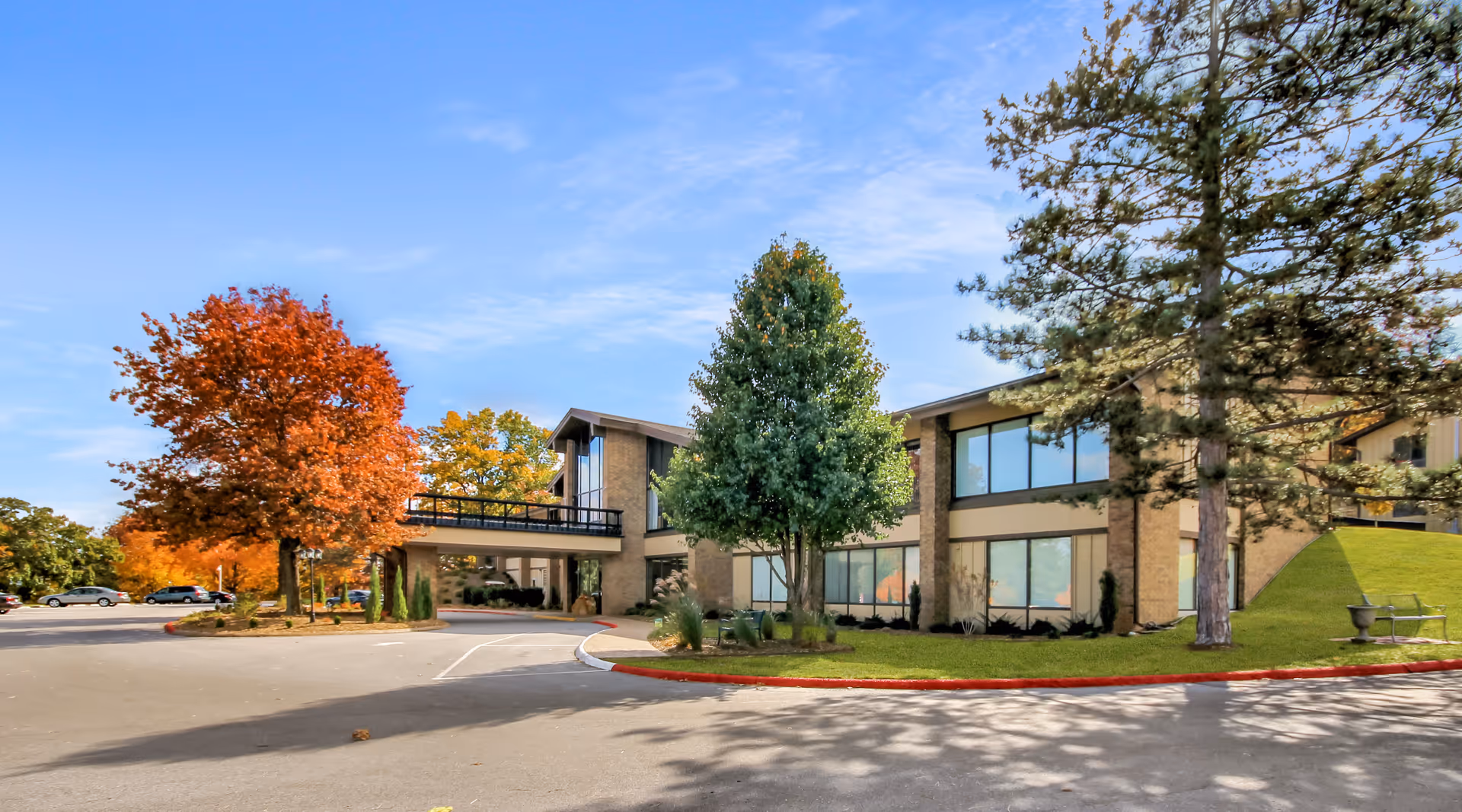 Exterior view of a two-story senior living facility building with large windows, surrounded by trees with autumn foliage and a clear blue sky. There is a driveway and parking area in front of the building.