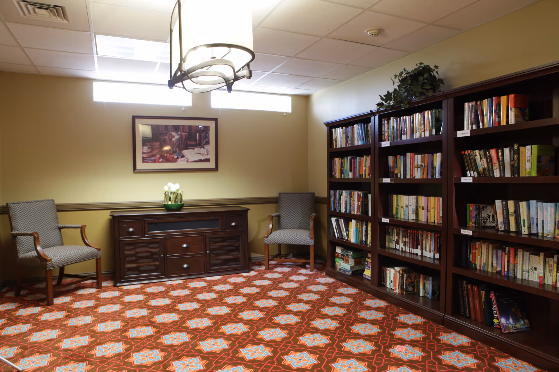A cozy reading room with patterned carpet, two upholstered armchairs, a wooden cabinet with a flower pot on top, and three large bookshelves filled with books. A framed picture hangs on the wall above the cabinet, and a ceiling light fixture illuminates the room.