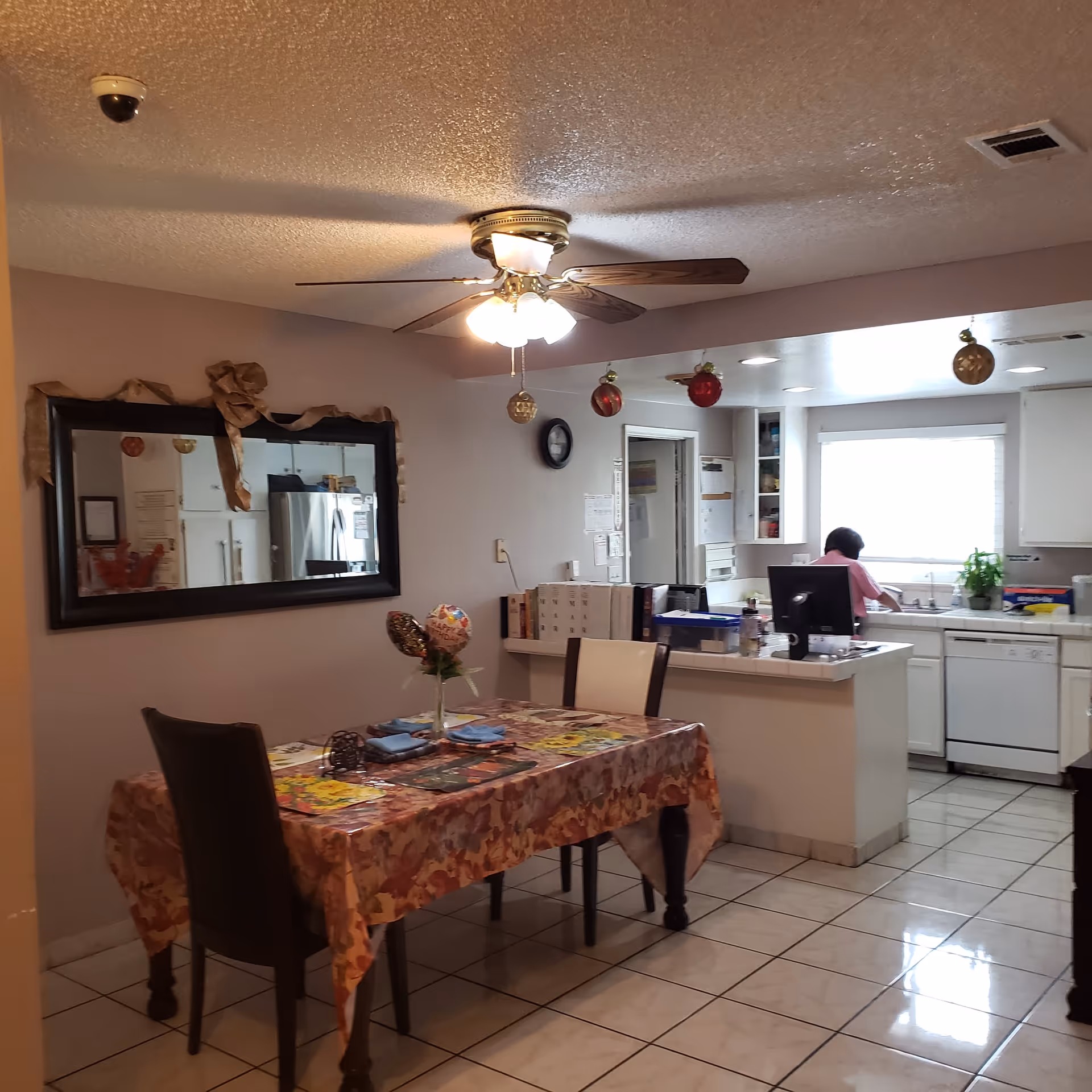 Interior view of a dining area and kitchen in a senior living facility. The dining table is covered with a floral tablecloth and set with placemats and napkins. Above the table, a ceiling fan with lights is turned on. A large mirror with a decorative ribbon is mounted on the wall. In the background, a person is standing at the kitchen sink, which is surrounded by white cabinets and appliances. Festive ornaments hang from the ceiling near the kitchen area.