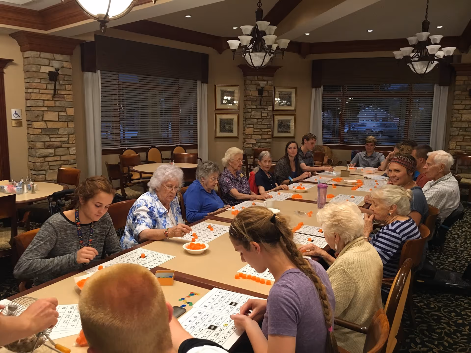A group of residents and visitors seated around a long table playing bingo in a community dining/activity room.