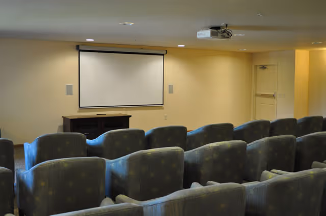 Small community media room with rows of upholstered chairs facing a wall-mounted projection screen and ceiling projector.