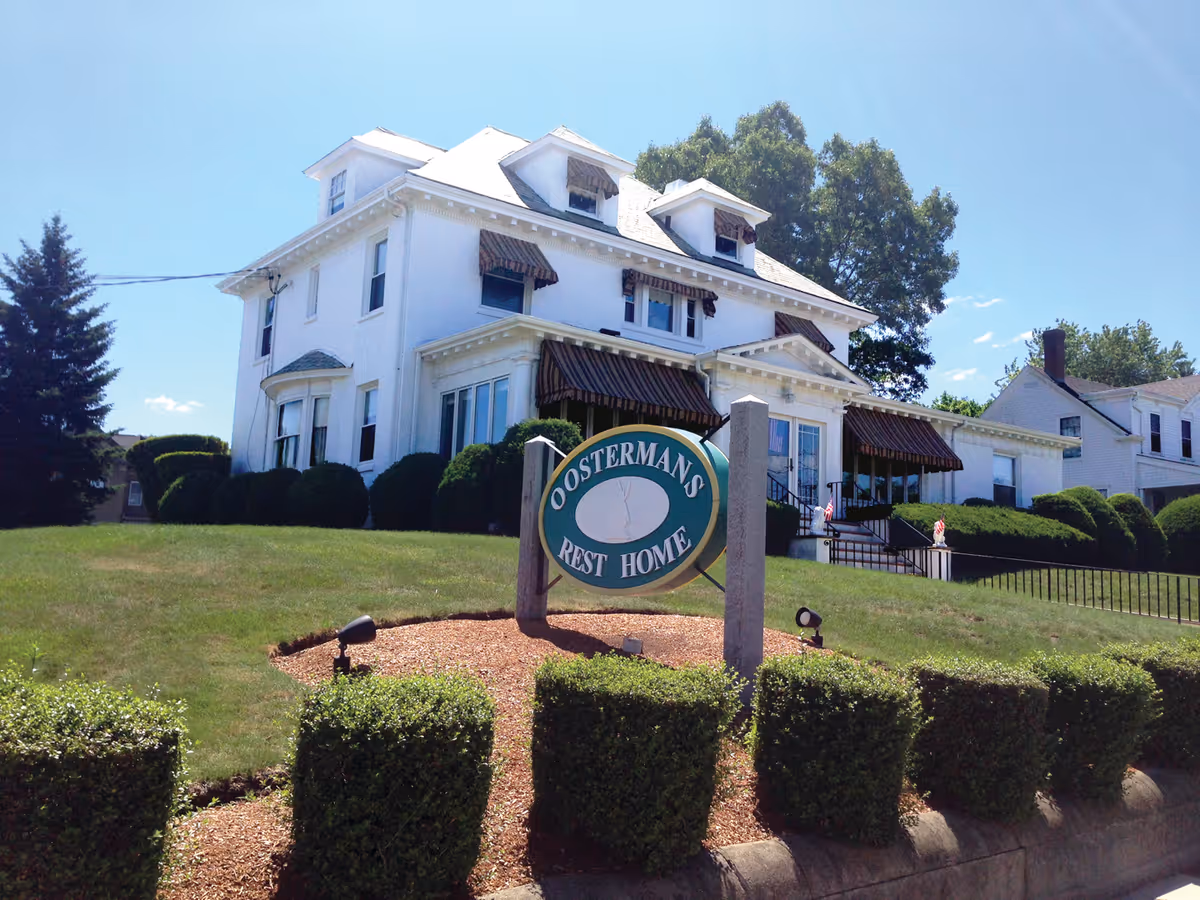 A large white multi-story house with brown striped awnings over the windows and entrance, surrounded by neatly trimmed bushes and a green lawn under a clear blue sky. In front of the house is a sign that reads 'Oosterman's Rest Home'.