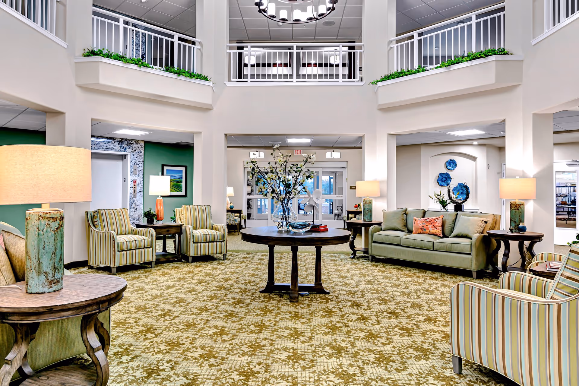Spacious and well-lit living room area in a senior living facility with patterned carpet, a round wooden table with a vase of flowers in the center, multiple armchairs with striped upholstery, a green sofa with cushions, side tables with lamps, and a balcony railing above with greenery.