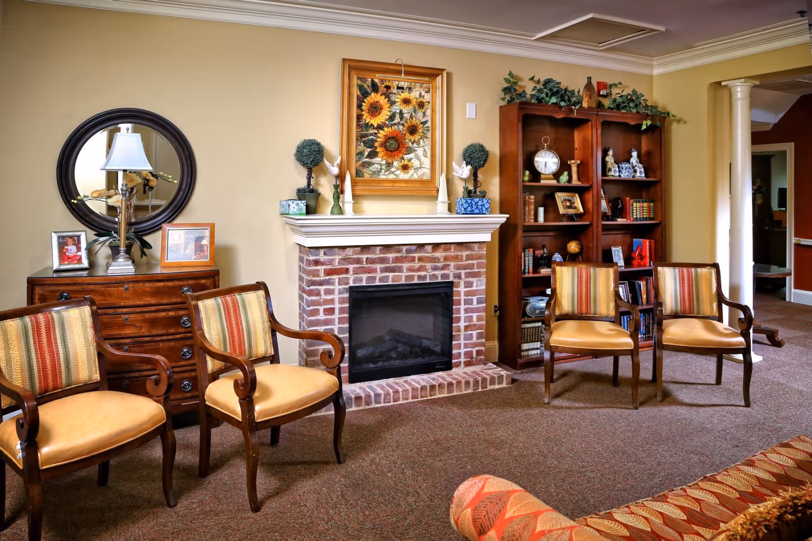 Cozy common room with a brick fireplace, wooden chairs, a bookshelf and decorative accents.