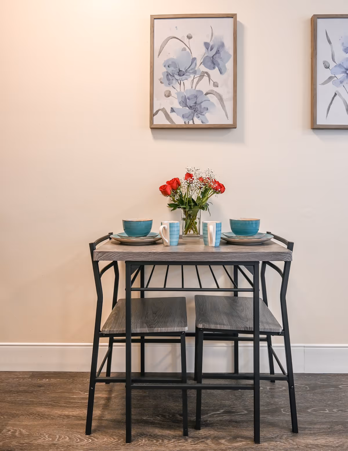 A small dining table set for two with two chairs, featuring teal bowls, striped mugs, and a vase with red roses and white baby's breath flowers. Two framed floral paintings hang on the beige wall behind the table.