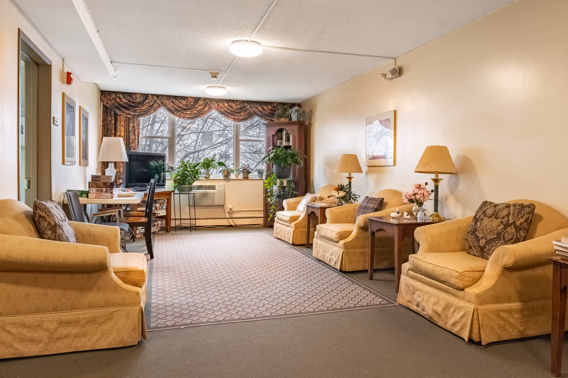 A cozy senior living common area with several beige upholstered armchairs arranged along the walls, each accompanied by side tables with lamps and floral decorations. A carpeted floor leads to a window with patterned curtains, and there are several potted plants on a table and a stand near the window. A small TV is placed on a wooden stand next to a table with chairs and board games stacked on it.