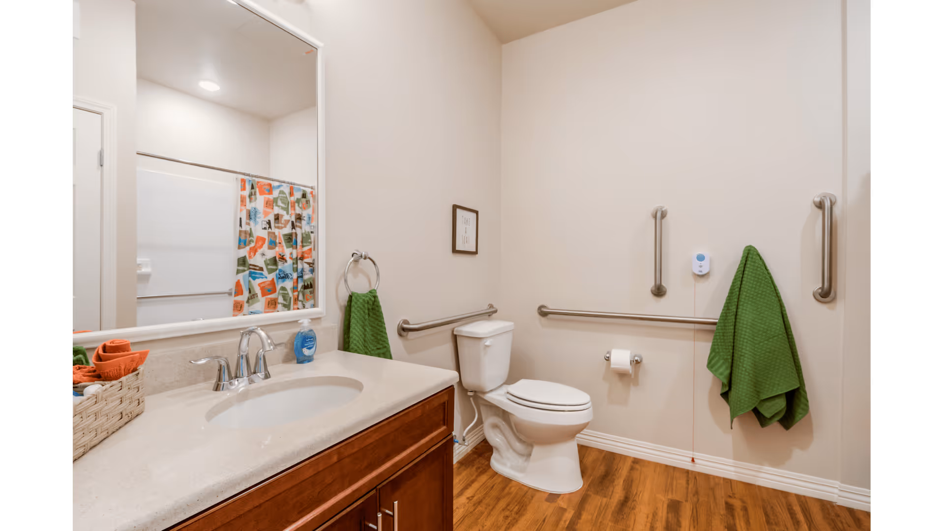A clean and accessible bathroom with a white toilet, a sink with a countertop and wooden cabinet, a large mirror above the sink, grab bars on the walls, green towels hanging on towel rings and hooks, and a shower with a colorful curtain in the background.