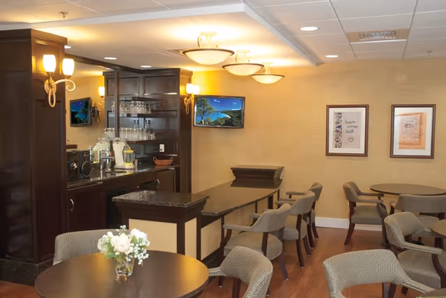 Interior view of a dining area in a senior living facility with round tables and cushioned chairs. A small bar area with glassware and a countertop is visible, along with warm lighting fixtures on the ceiling and walls. Two framed pictures hang on the beige wall, and a small vase with white flowers is on one of the tables.