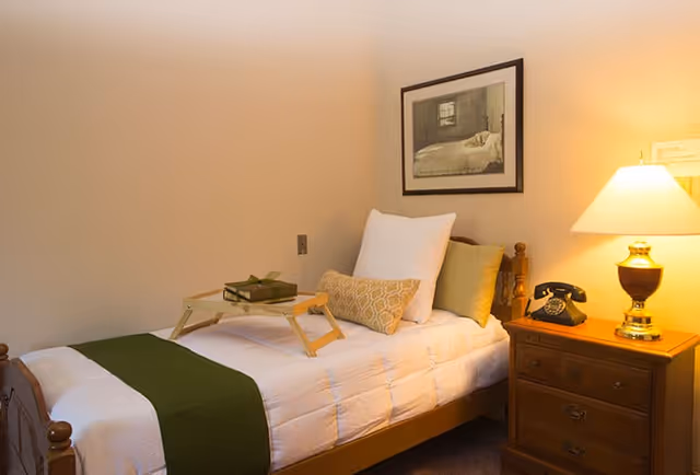 A cozy senior living bedroom with a single wooden bed dressed in white bedding and a green runner. The bed has multiple pillows and a small wooden tray with a book and glasses on it. Next to the bed is a wooden nightstand with an old-fashioned black rotary phone and a lit table lamp. A framed picture hangs on the beige wall above the bed.
