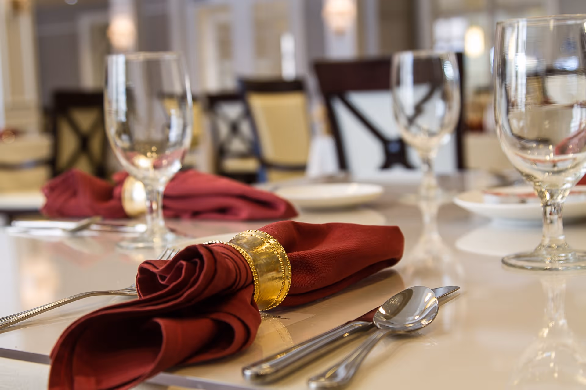 Close-up view of a dining table set with red cloth napkins held by gold napkin rings, clear water glasses, silverware including forks and spoons, and white plates in an elegant dining room setting.