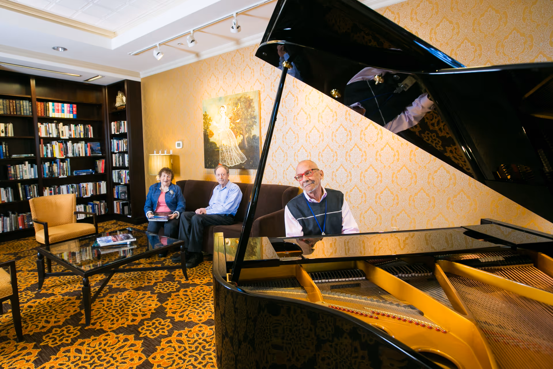 A grand piano in a lounge with a man playing while two older adults sit on a sofa near a bookshelf.