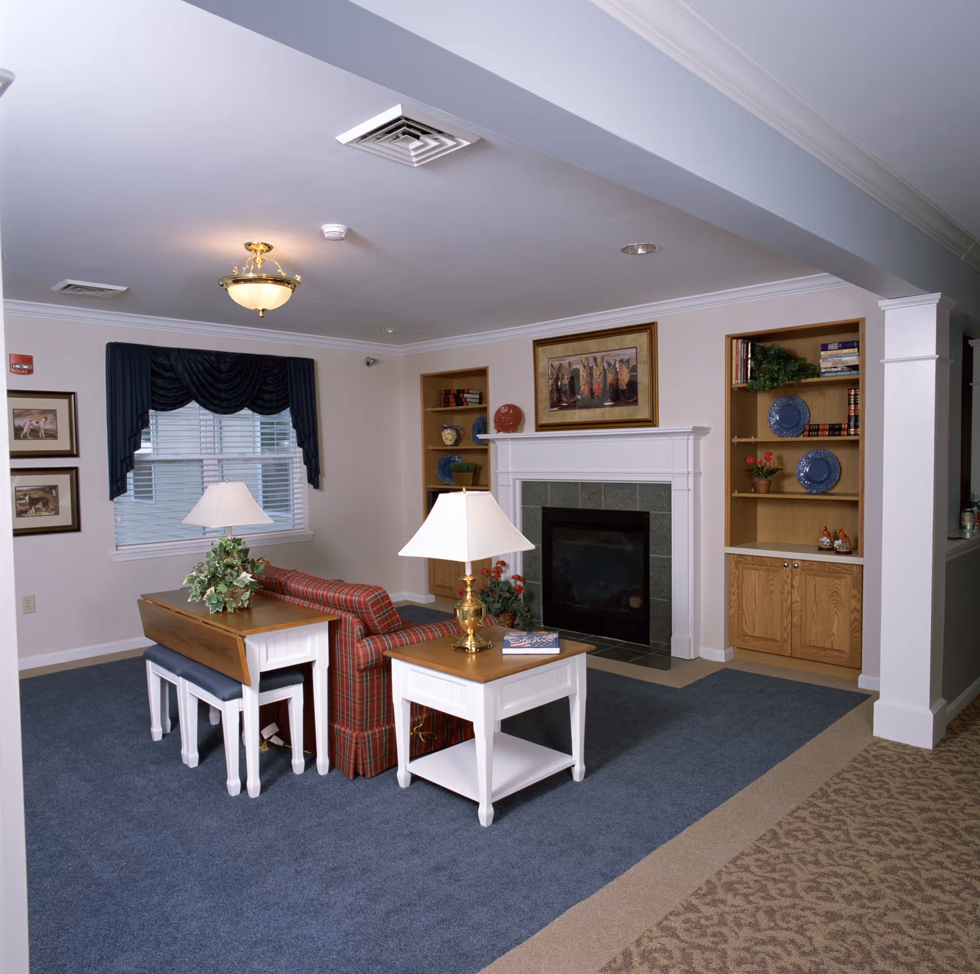A cozy living room area with a plaid sofa, two white side tables with lamps, a fireplace with a framed painting above it, built-in wooden shelves with decorative items and books, a window with dark blue curtains, and carpeted flooring.