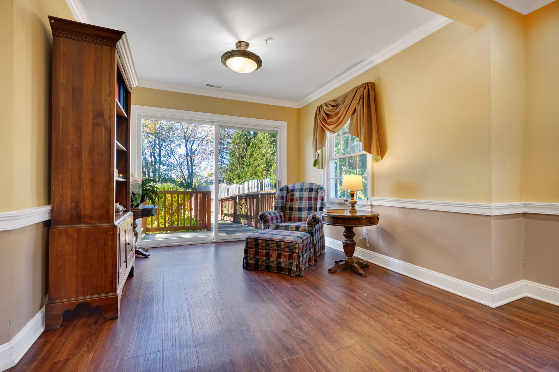 A cozy living room area with wooden flooring, a plaid armchair with matching ottoman, a round wooden side table with a lamp, a large wooden bookshelf, and a sliding glass door leading to an outdoor deck with trees visible outside.