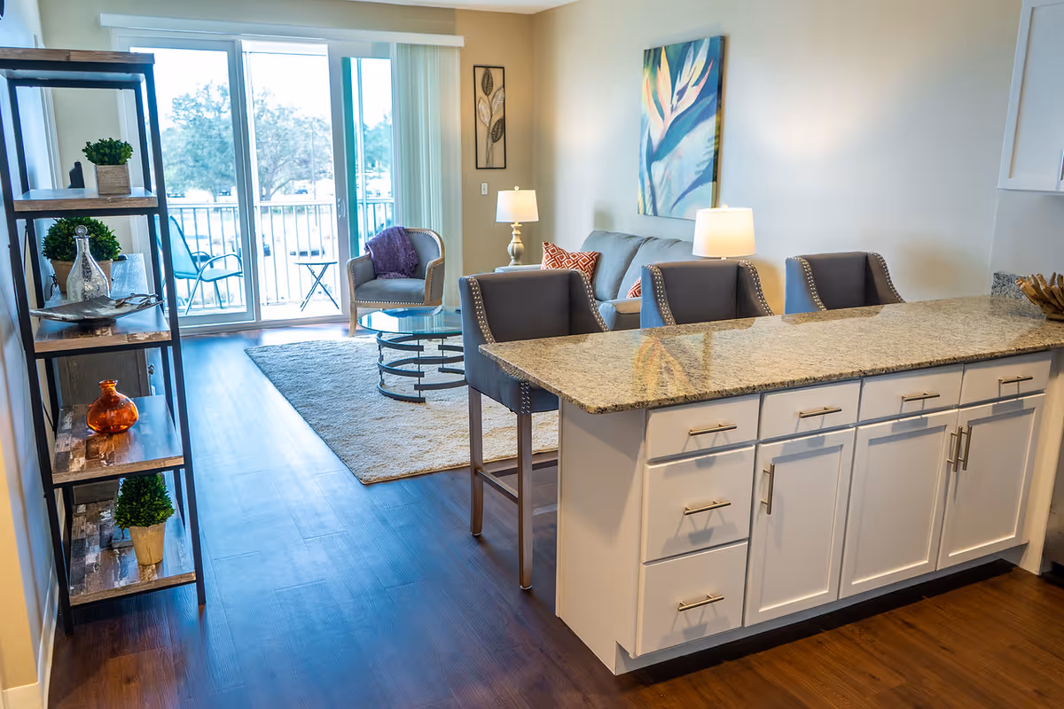 A bright and modern living area with a kitchen island featuring a granite countertop and three upholstered bar stools. The living room has a gray sofa with patterned pillows, a glass coffee table on a beige rug, and a single armchair near sliding glass doors that lead to a balcony with outdoor seating. Decorative plants and artwork adorn the walls and shelves.