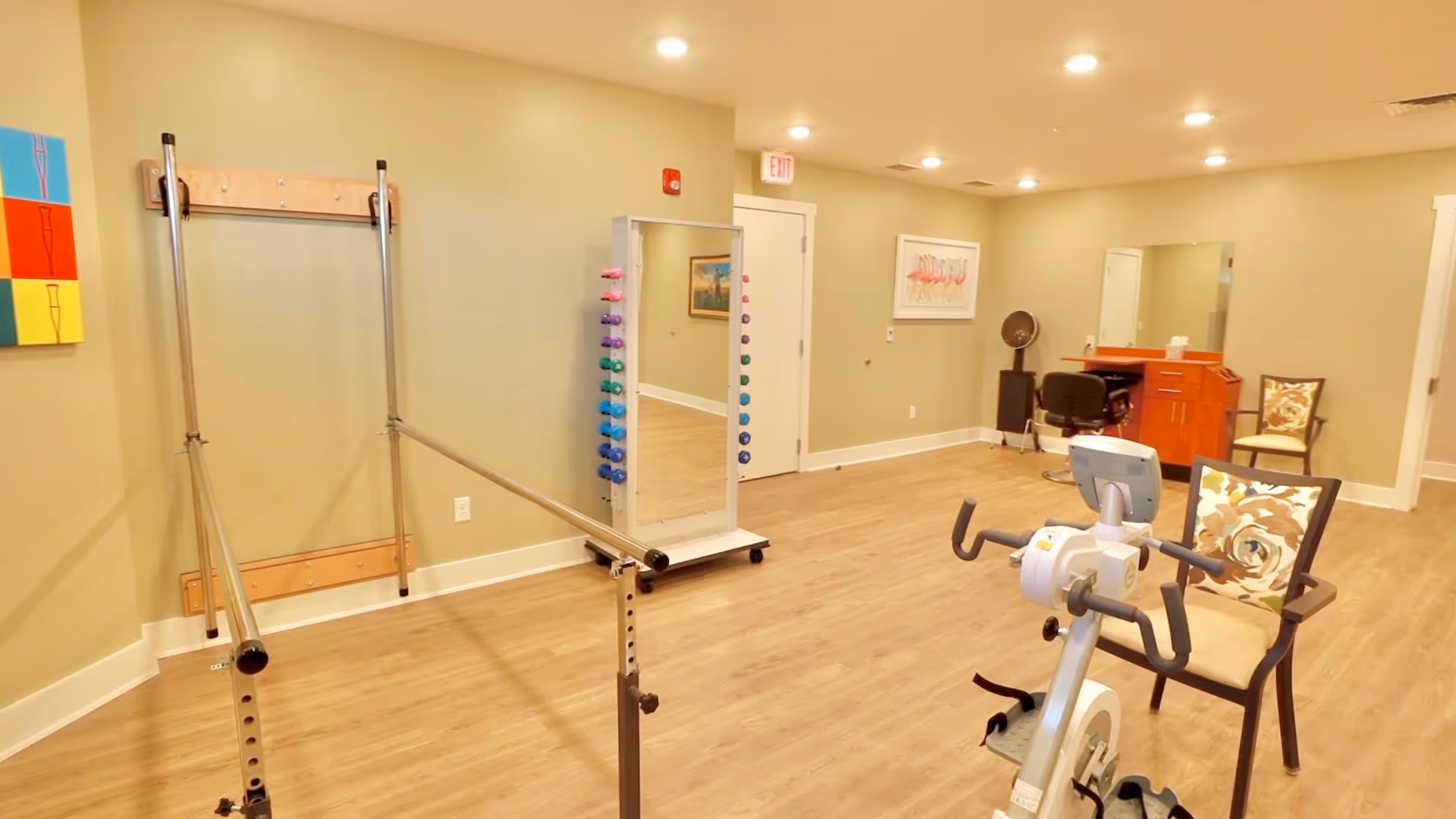 A well-lit exercise room with wooden flooring and light green walls. The room contains parallel bars for walking exercises, a stationary exercise bike, a full-length mirror with colorful dumbbells attached on the sides, a small desk with a chair, and two cushioned chairs with floral-patterned pillows. There is a colorful artwork on the left wall and a framed picture of flamingos on the far wall.