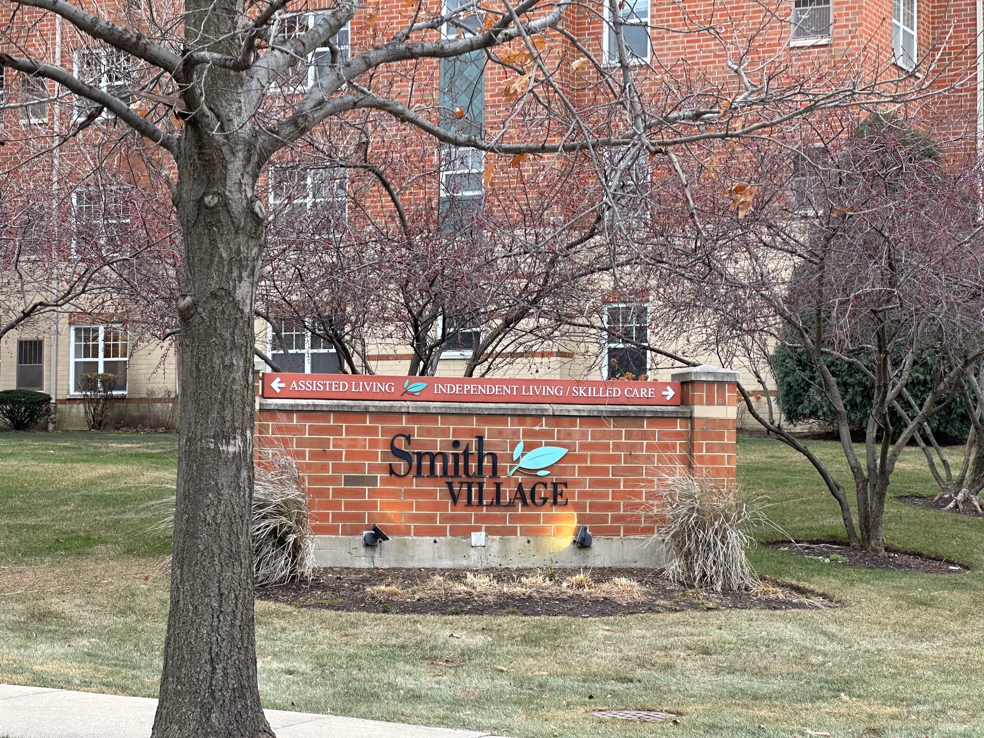 A brick sign for Smith Village in front of a building with leafless trees and grass surrounding it. The sign includes directional arrows for Assisted Living to the left and Independent Living / Skilled Care to the right.