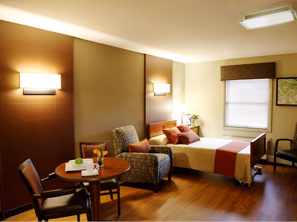 A cozy senior living bedroom with a single bed adorned with orange and beige bedding and pillows. Next to the bed is a wooden nightstand with a lamp and a small vase of flowers. There is a patterned armchair and a small round wooden table with two chairs, one holding a green apple and a cup. The room has warm lighting from wall sconces and a ceiling light, with a window covered by blinds and a brown valance. The floor is wooden, and the walls are painted in neutral tones.