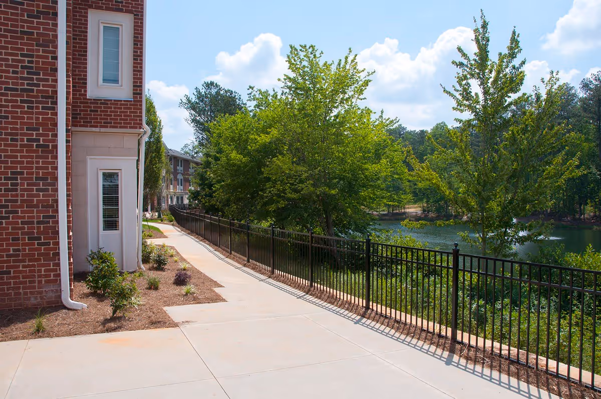 A paved walkway alongside a brick building with windows, bordered by a black metal fence. On the other side of the fence, there are green trees and a body of water under a partly cloudy sky.