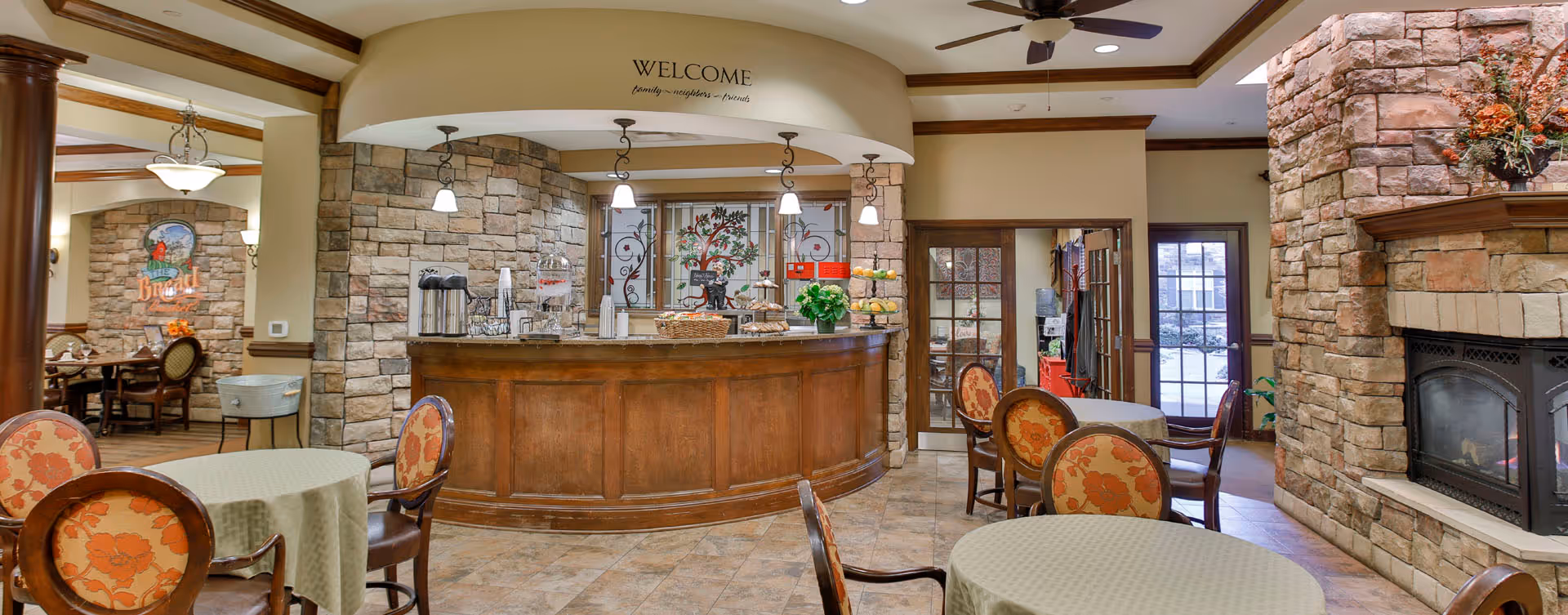 A cozy dining area in a senior living facility featuring round tables with floral upholstered chairs and green tablecloths. There is a wooden counter with hanging pendant lights above it, decorated with a water dispenser, coffee urns, baskets of snacks, and a flower arrangement. The walls are made of stone and beige paint, with a fireplace on the right side and a welcoming sign above the counter that reads 'WELCOME family ~ neighbors ~ friends'. The room has a warm and inviting atmosphere with wooden ceiling beams and a ceiling fan.