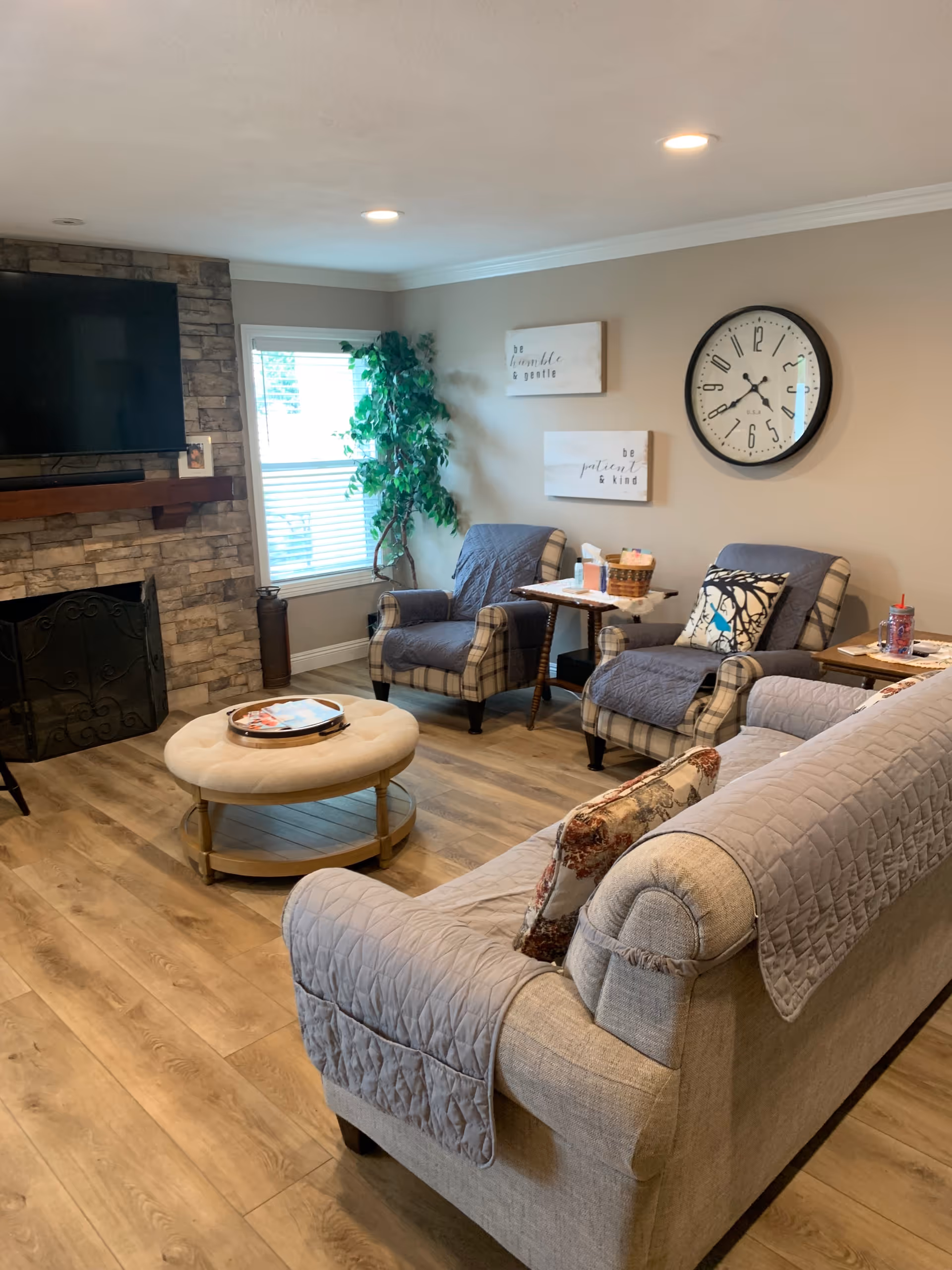 A cozy living room with a beige sofa covered with a quilted throw, two plaid armchairs with gray covers, a round beige ottoman coffee table, a stone fireplace with a mounted flat-screen TV above it, a large wall clock, two framed inspirational quotes on the wall, a window with blinds, and a tall green plant in the corner.