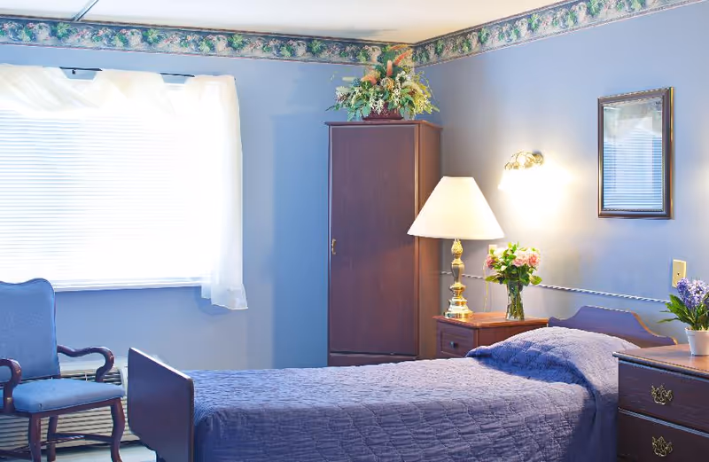 A neatly made single bed with a blue quilt in a softly lit bedroom. The room features a wooden nightstand with a lamp and a vase of flowers, a wooden wardrobe with a floral arrangement on top, a blue upholstered chair, and a window with white curtains allowing natural light to enter. The walls are painted blue with a floral border near the ceiling.
