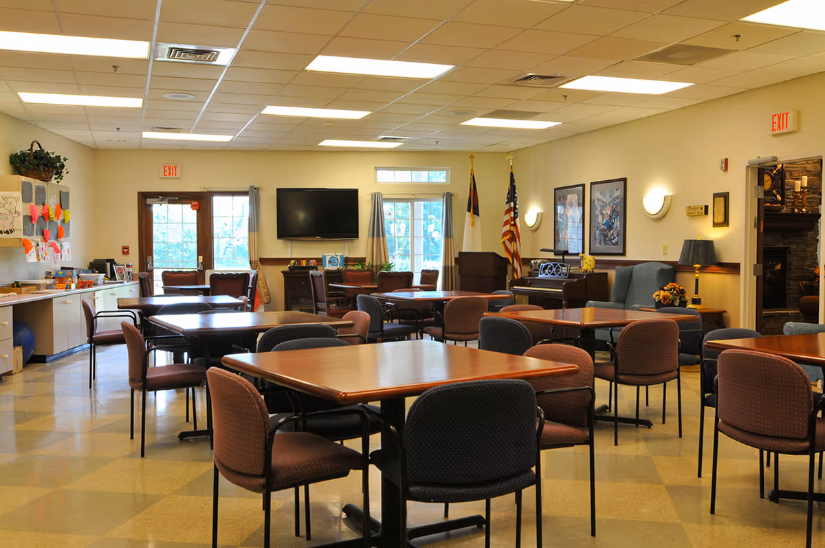 A spacious common room with multiple square tables and chairs arranged throughout. The room has large windows with curtains, a wall-mounted TV, and two flags near a podium. There is a piano and comfortable seating area with armchairs and framed artwork on the walls. The room is well-lit with ceiling lights and has a tiled floor.