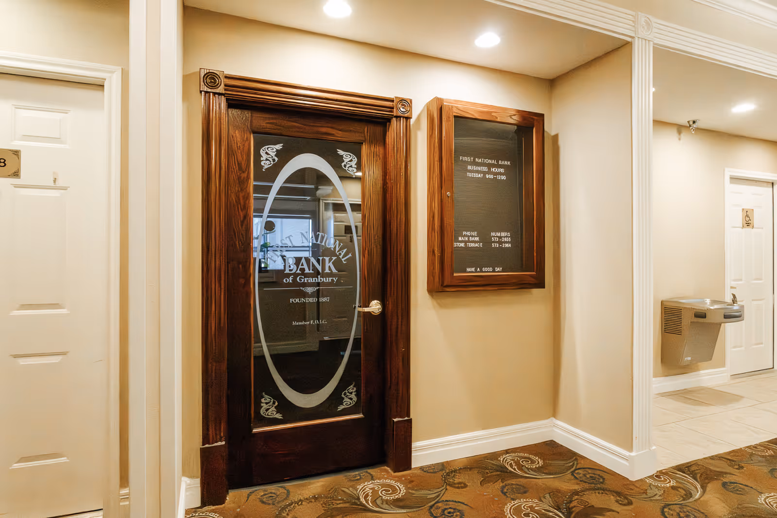 Interior hallway with beige walls and patterned carpet. A wooden door with frosted glass displays the text 'First National Bank of Granbury Founded 1887'. Next to the door is a wooden-framed bulletin board with information about the bank's business hours and phone numbers. A water fountain is mounted on the wall further down the hallway, near a white door with an accessibility sign.