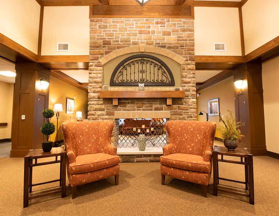 A cozy seating area in a senior living facility featuring two orange patterned armchairs facing a stone fireplace with a decorative wrought iron grate. Each chair is accompanied by a wooden side table, one with a potted plant and the other with a vase of flowers. The room has warm lighting, wooden beams, and beige walls.