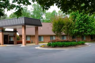 Exterior view of B'nai B'rith Homecrest House showing the entrance with a covered driveway supported by brick columns, surrounded by trees and shrubs.