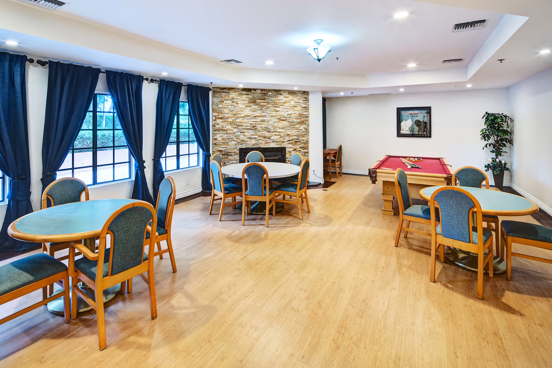 A spacious common room with multiple round tables and chairs, a pool table with a red felt surface, large windows with dark blue curtains, a stone accent wall with a fireplace, and a framed picture on the wall.