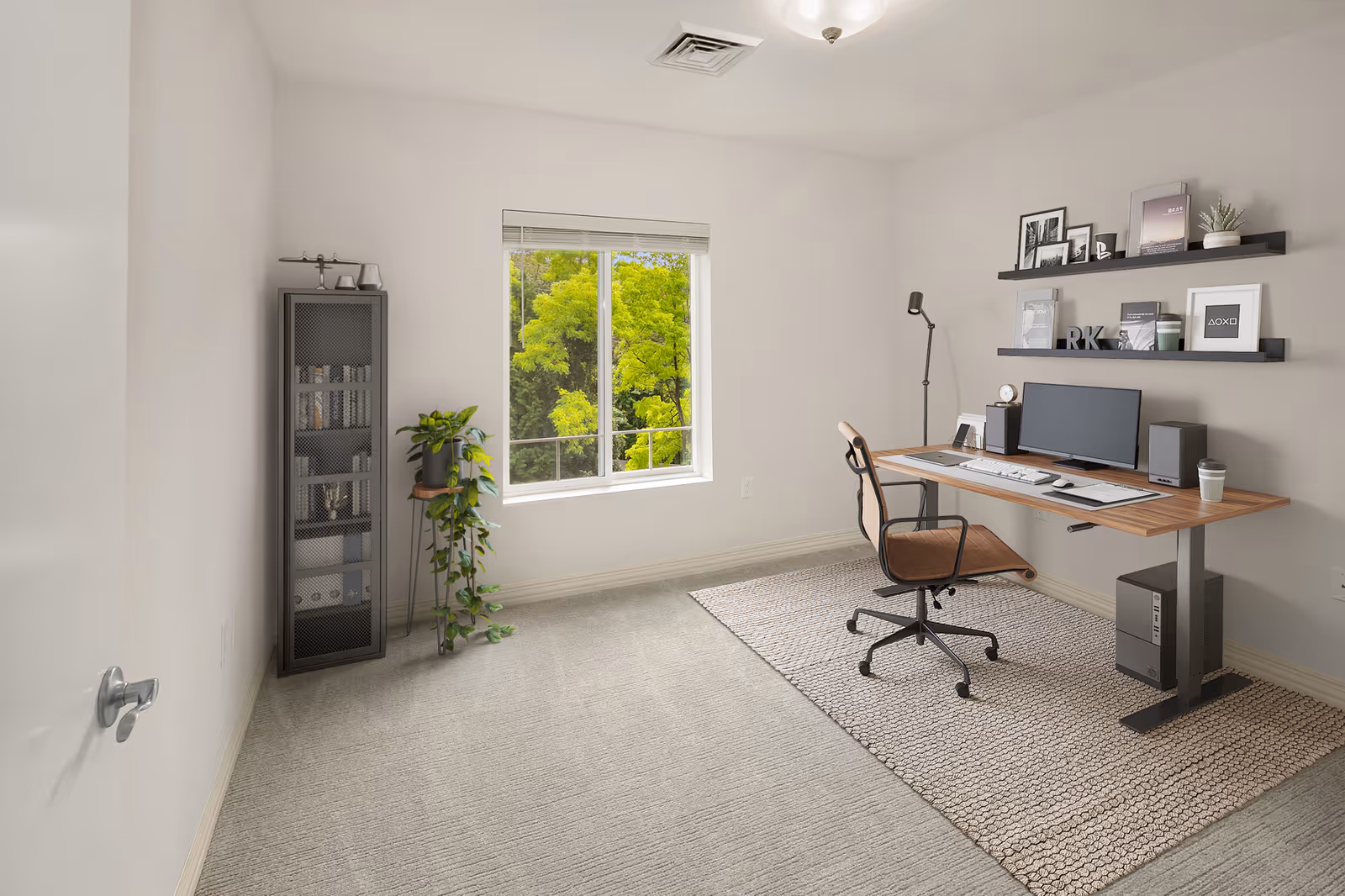 A bright, minimalist home office with a large window showing green trees outside. The room features a wooden desk with a computer, keyboard, mouse, speakers, and a cup. A brown office chair is positioned at the desk on a textured rug. Two black floating shelves on the wall hold framed pictures, books, and decorative items. A tall black cabinet with glass doors and a potted plant on a stand are placed near the window. The walls and carpet are light-colored, creating a clean and airy atmosphere.