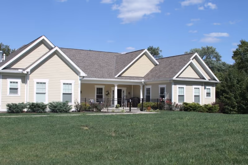 Exterior view of a single-story beige senior living facility building with white trim, multiple windows, a covered porch with columns, and a well-maintained green lawn under a blue sky with some clouds.