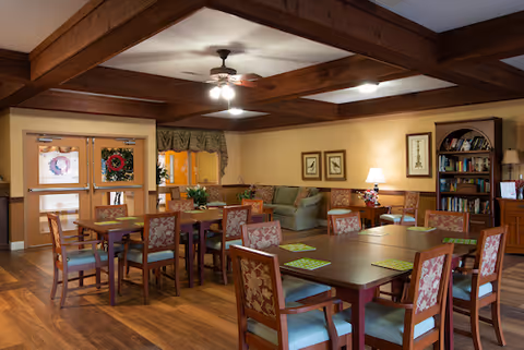 A cozy common area with wooden beams on the ceiling, multiple tables and chairs with floral upholstery, a green sofa, a bookshelf filled with books, framed artwork on the walls, a ceiling fan with lights, and double doors decorated with holiday wreaths.